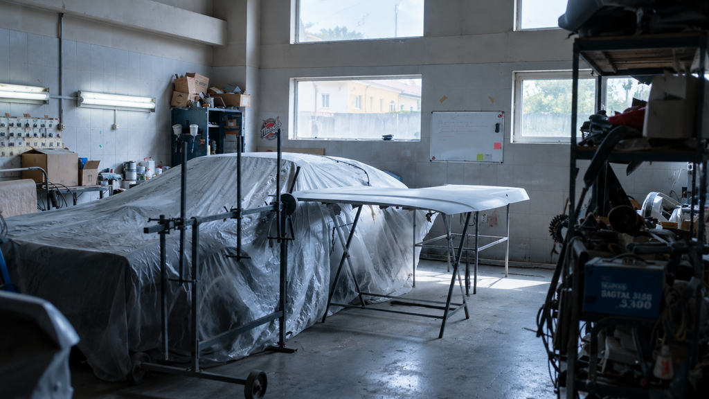 A car under a plastic cover in a dim workshop, with a hood on a stand beside it